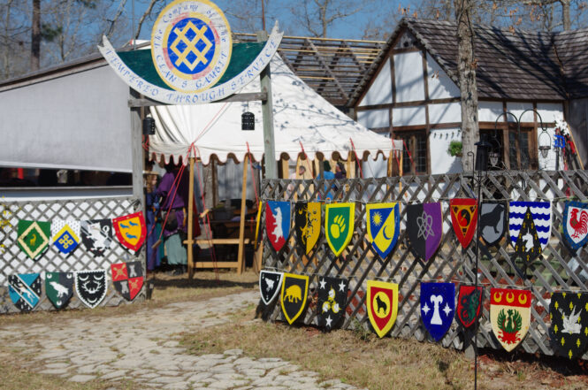 A gate and fence featuring many brightly colored heraldic shields with tents and houses behind the gate.